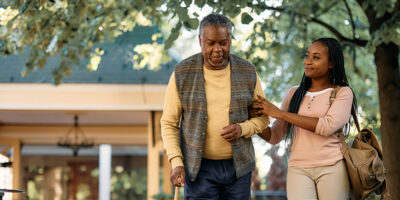 Black senior man with walking cane and his daughter taking a walk through the park of a nursing home.